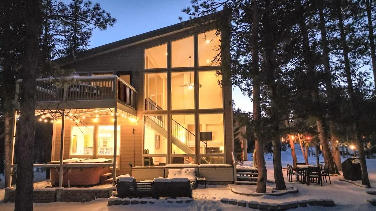 Two-story glass house at dusk with snow, patio seating and hot tub