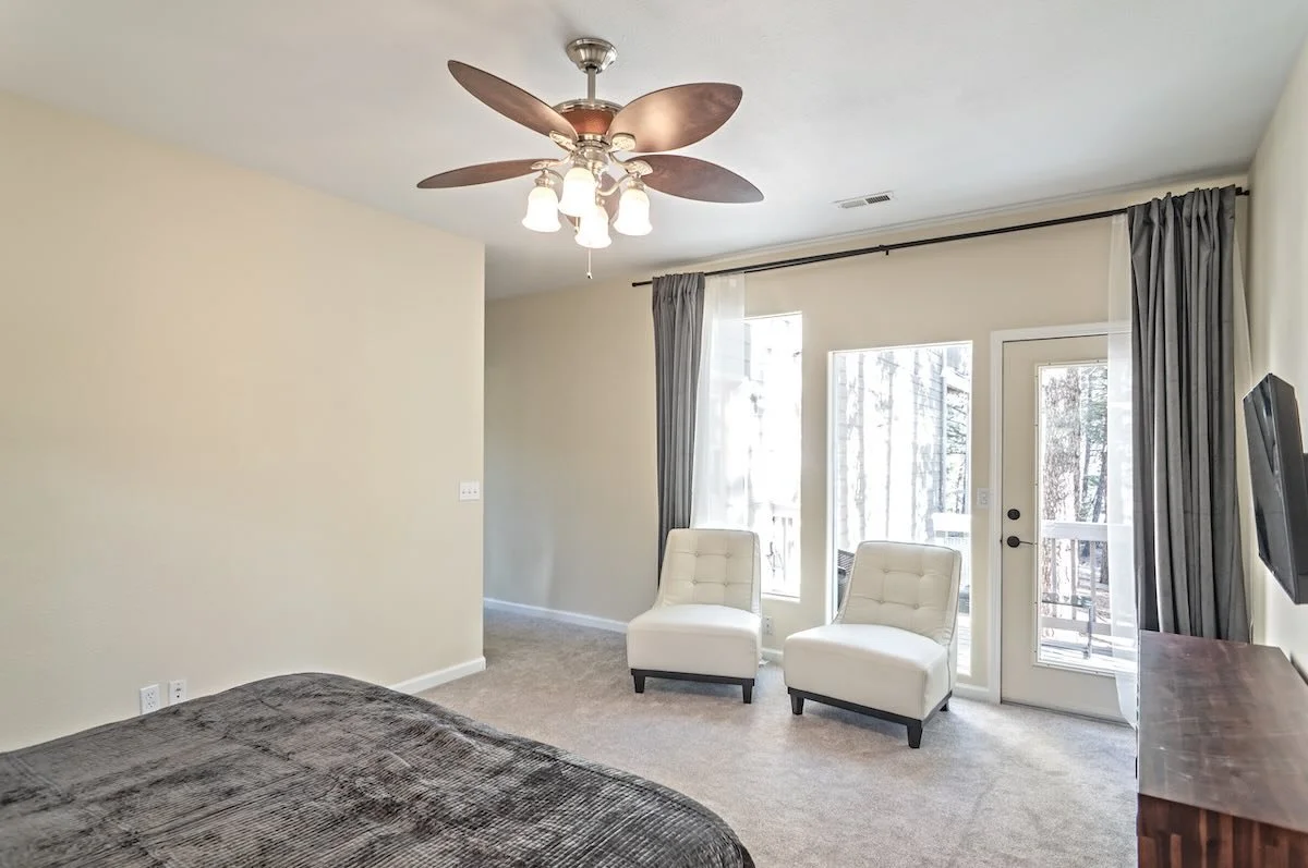 Primary bedroom with ceiling fan, two white chairs by the glass doors and a wall-mounted TV